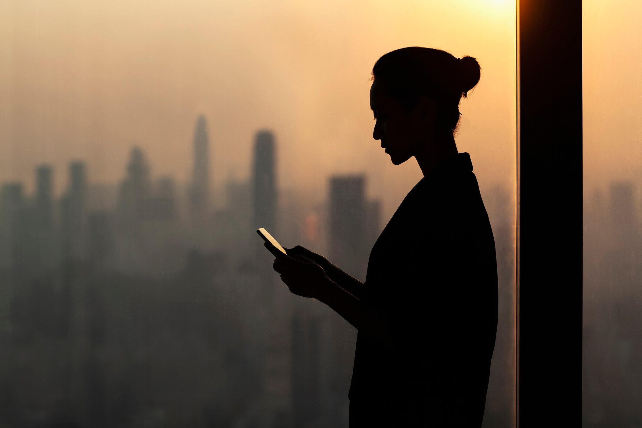 Silhouette of young woman using smartphone next to window with cityscape, Shenzhen, China